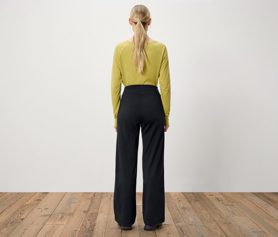 Vue de dos, une femme aux cheveux blonds attachés en queue de cheval porte un haut à manches longues vert olive et un pantalon noir à jambes larges.