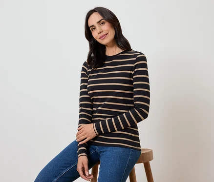 Une femme assise sur un tabouret porte un t-shirt rayé noir et beige à manches longues et un jean bleu.