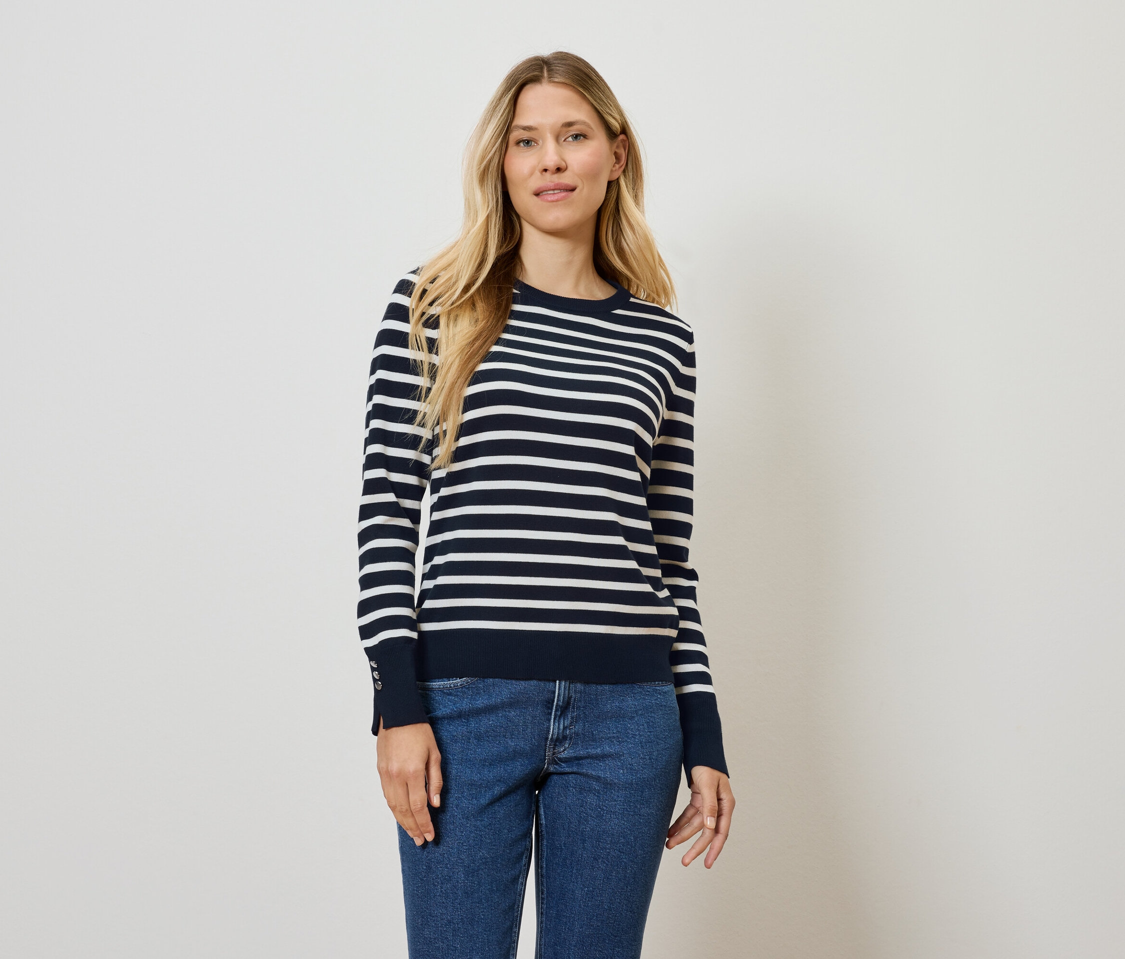 Une femme debout pose dans un pull rayé bleu et blanc et un jean.