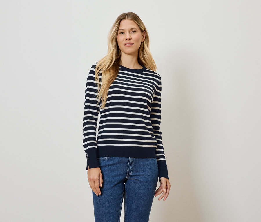 Une femme debout pose dans un pull rayé bleu et blanc et un jean.