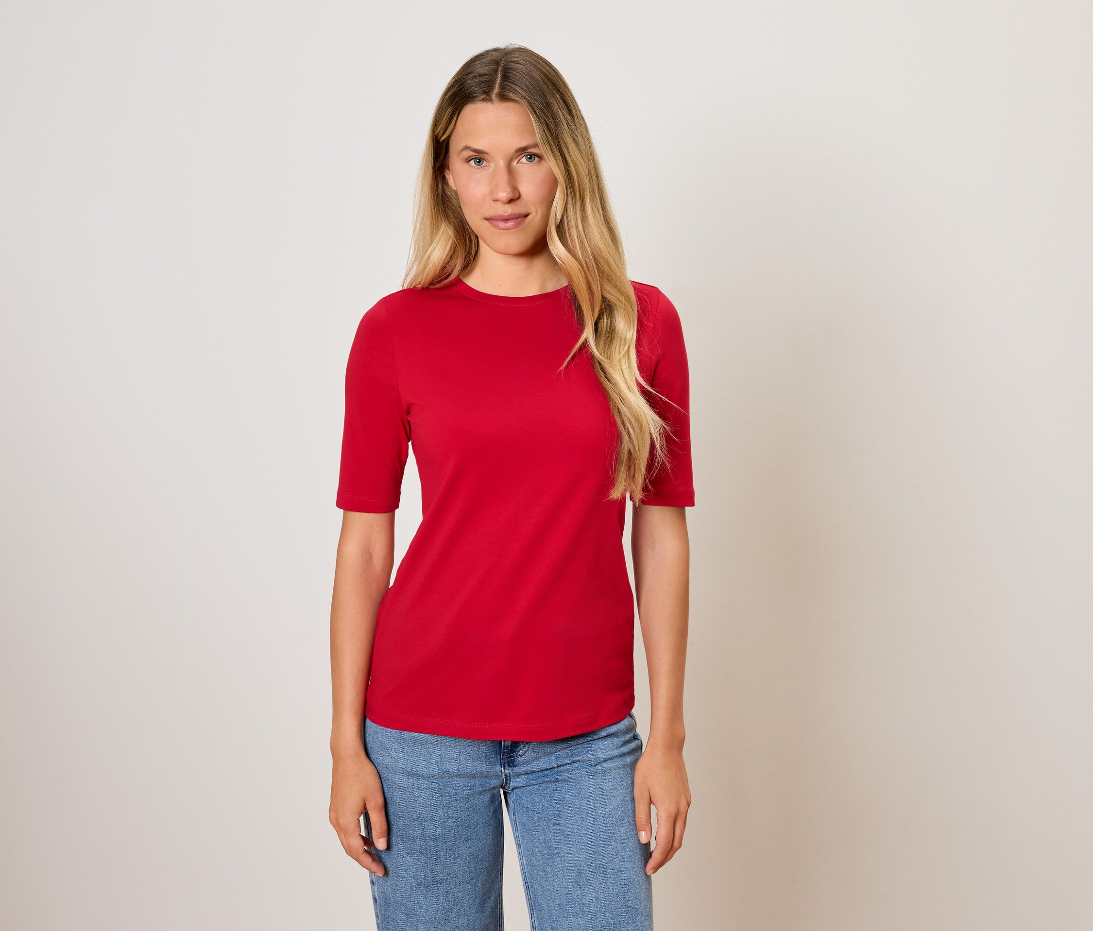 Femme aux longs cheveux blonds posant en t-shirt rouge et jean bleu.
