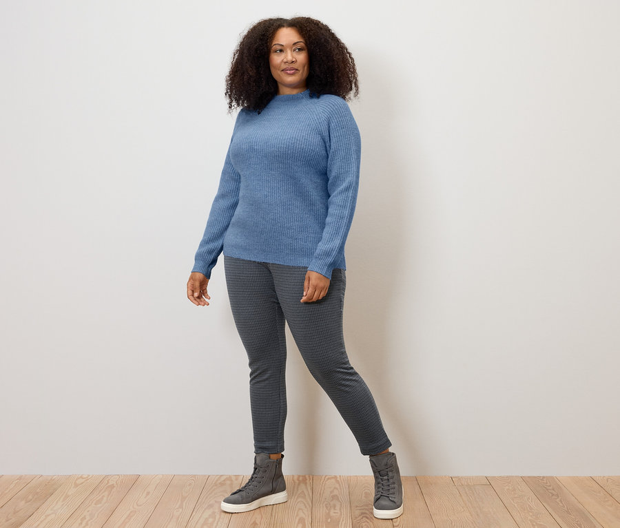 Une femme debout sur un plancher en bois portant un pull bleu et un pantalon en tissu à jacquard pied-de-poule gris.