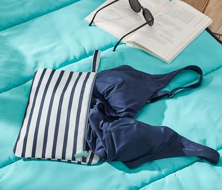 Sur une couverture bleue sont posés un sac pour affaires mouillées, un maillot de bain bleu, un livre et des lunettes de soleil.