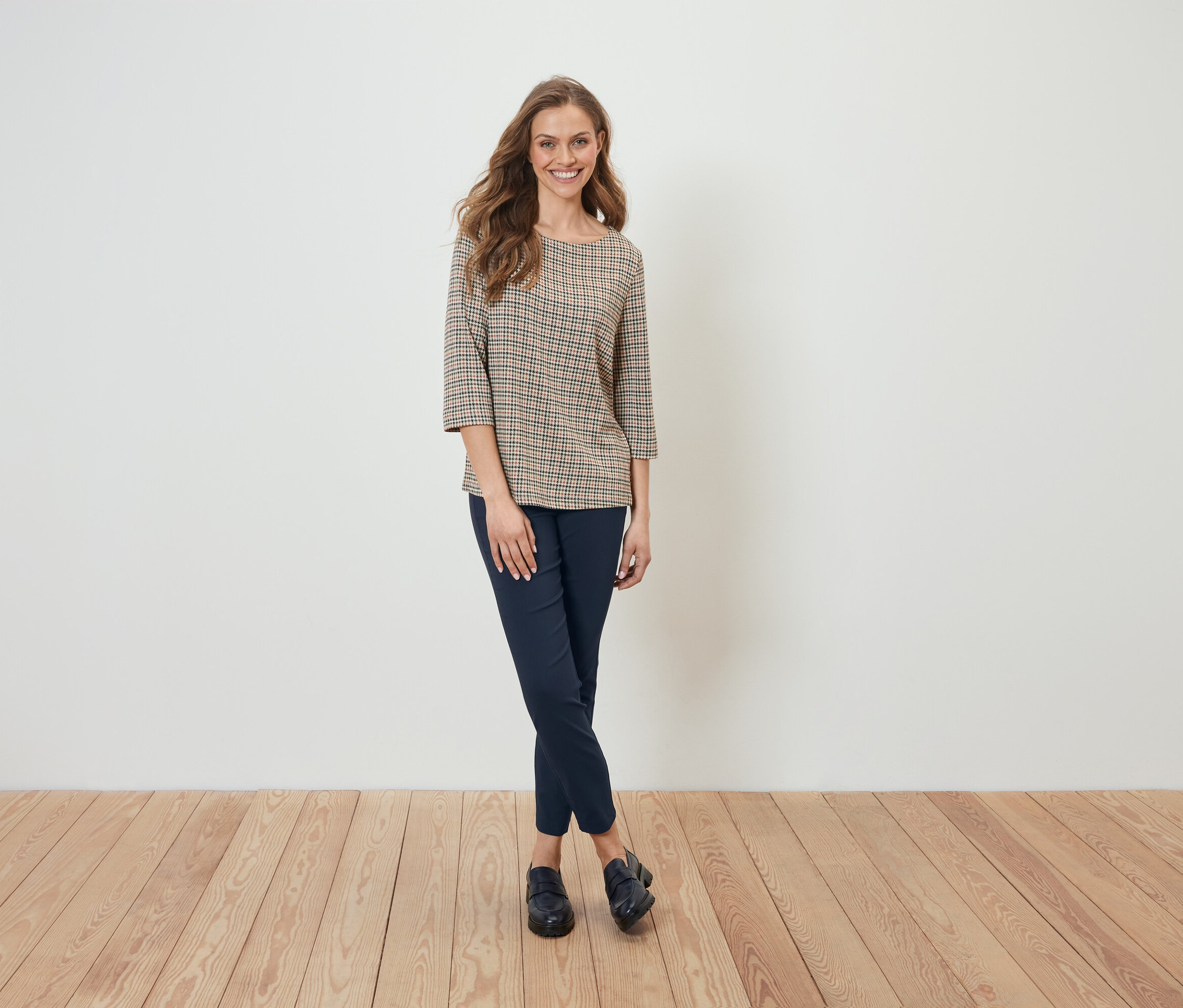 Une femme en chemisier à carreaux et pantalon bleu marine se tient devant un mur blanc.