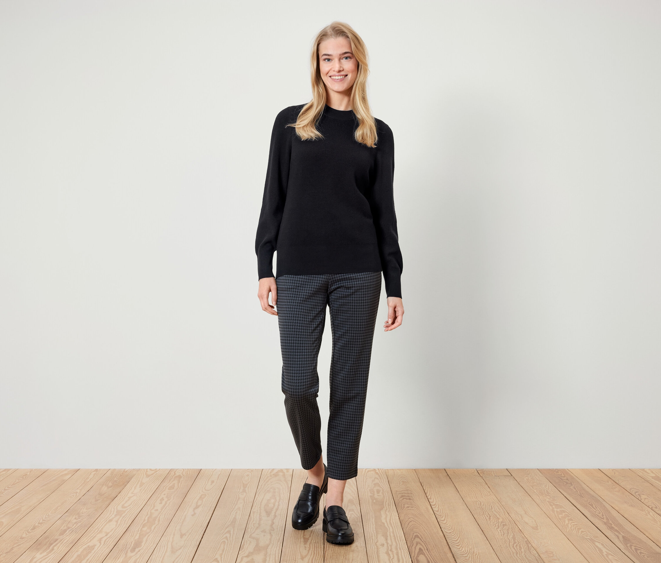 Une femme debout souriant, portant un pull noir, un pantalon à motifs et des mocassins noirs.