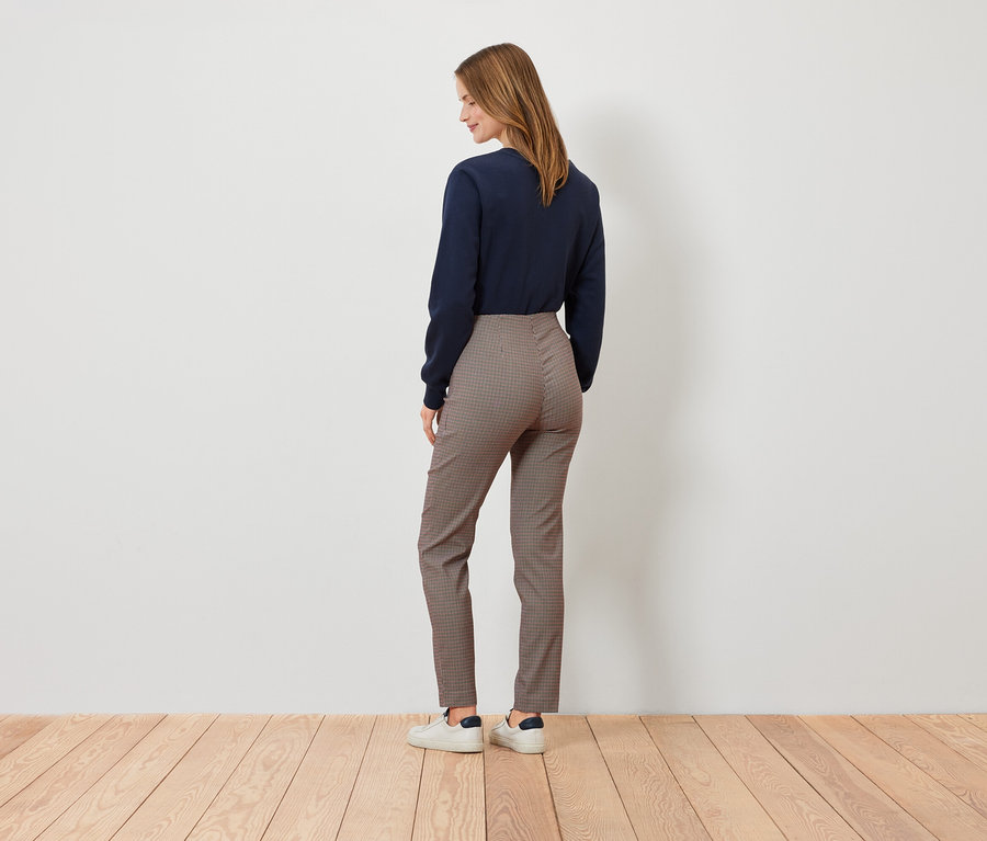 Une femme, vue de dos, porte un pull bleu marine, un pantalon à carreaux marron et des baskets blanches.