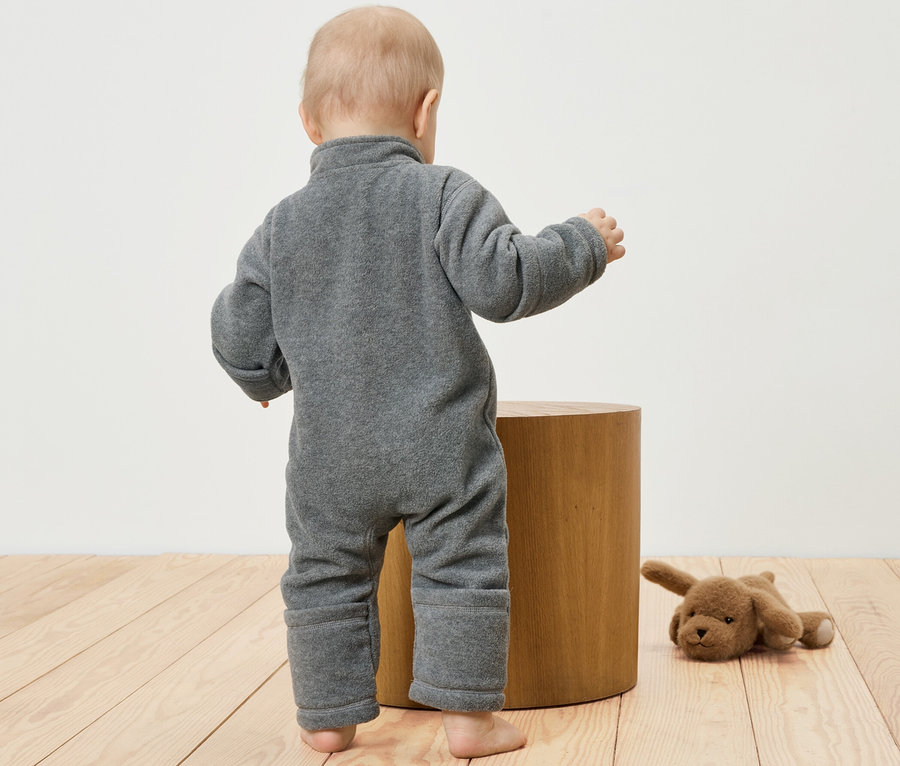 Un bébé dans une combinaison en polaire grise se tient à côté d'un tabouret en bois. Un chien en peluche est allongé à côté.