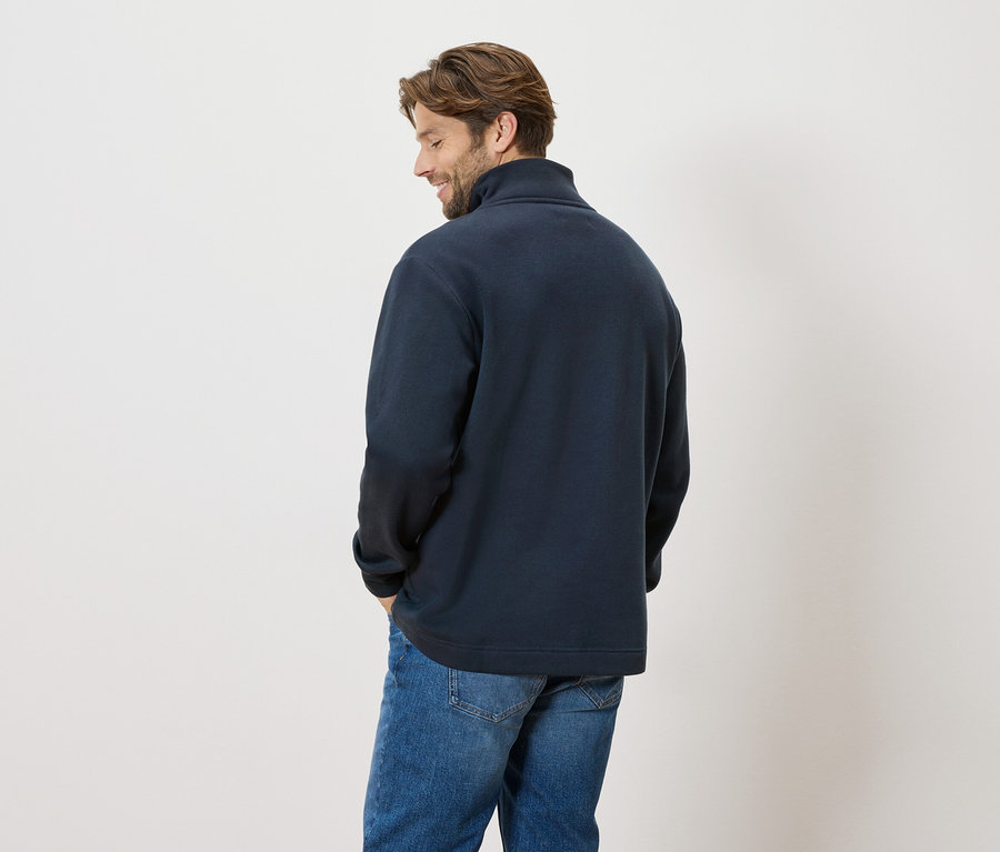 Un homme se tient dos à la caméra, il porte un pull bleu marine et un jean.