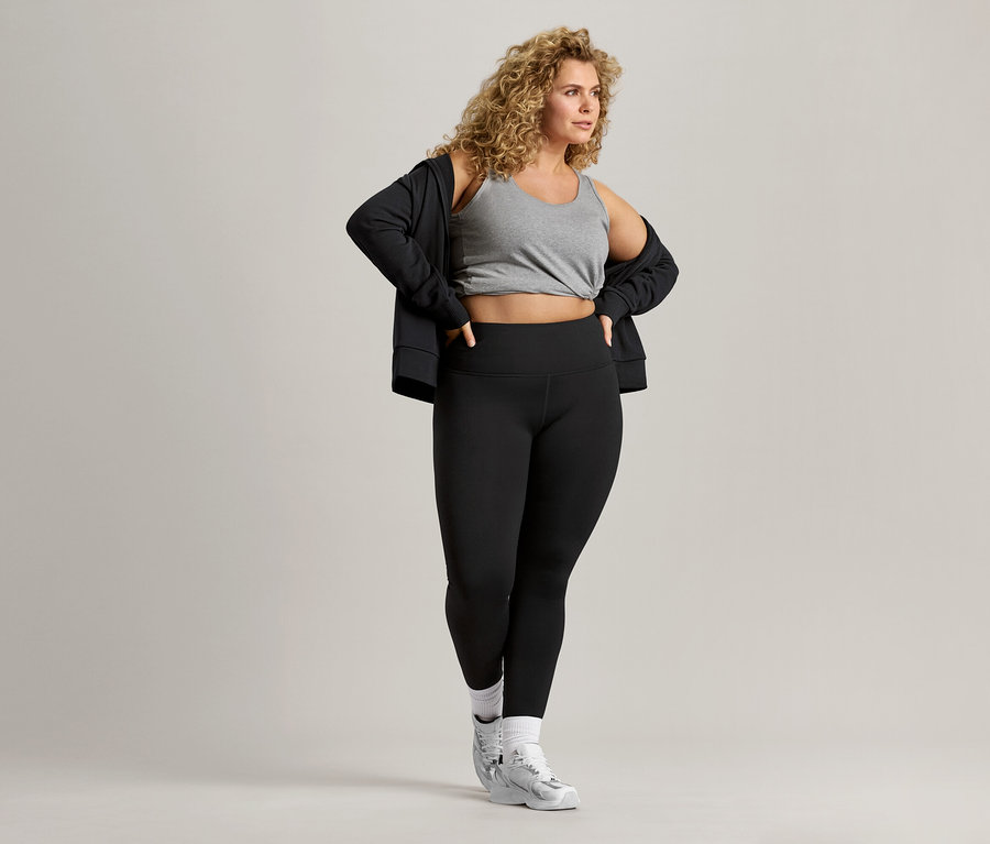 Une femme aux cheveux bouclés porte un débardeur gris, une veste noire et un legging noir.