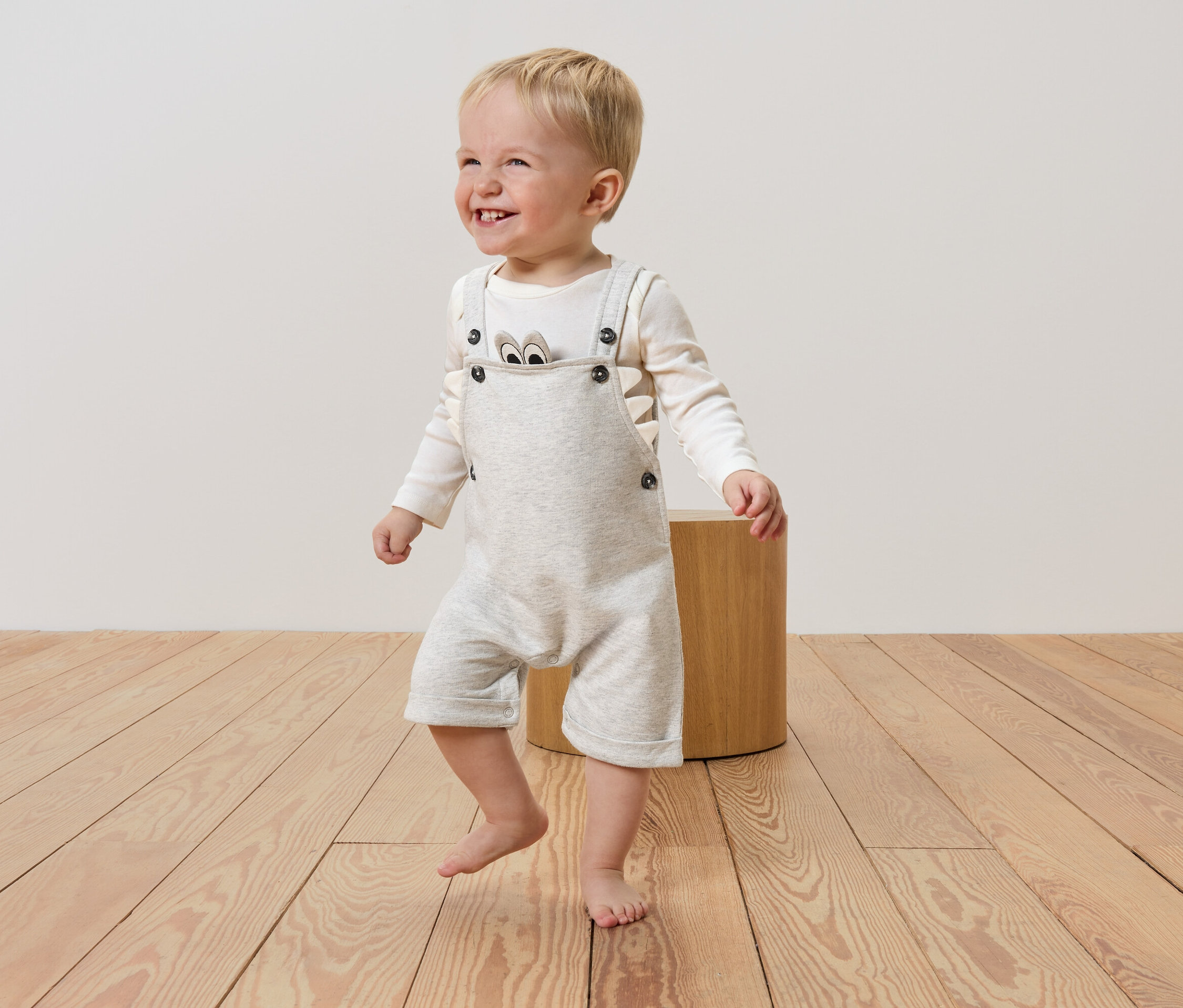 Un garçon blond souriant aux pieds nus porte une salopette gris clair boutonnée et un t-shirt à manches longues avec un imprimé d'ours, debout sur un plancher en bois à côté d'un tabouret en bois.