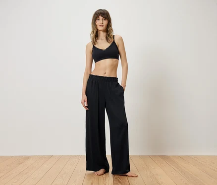 Femme debout pieds nus sur un plancher en bois, portant un soutien-gorge noir et un pantalon ample et fluide, mains dans les poches.