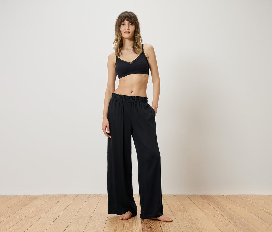 Femme debout pieds nus sur un plancher en bois, portant un soutien-gorge noir et un pantalon ample et fluide, mains dans les poches.
