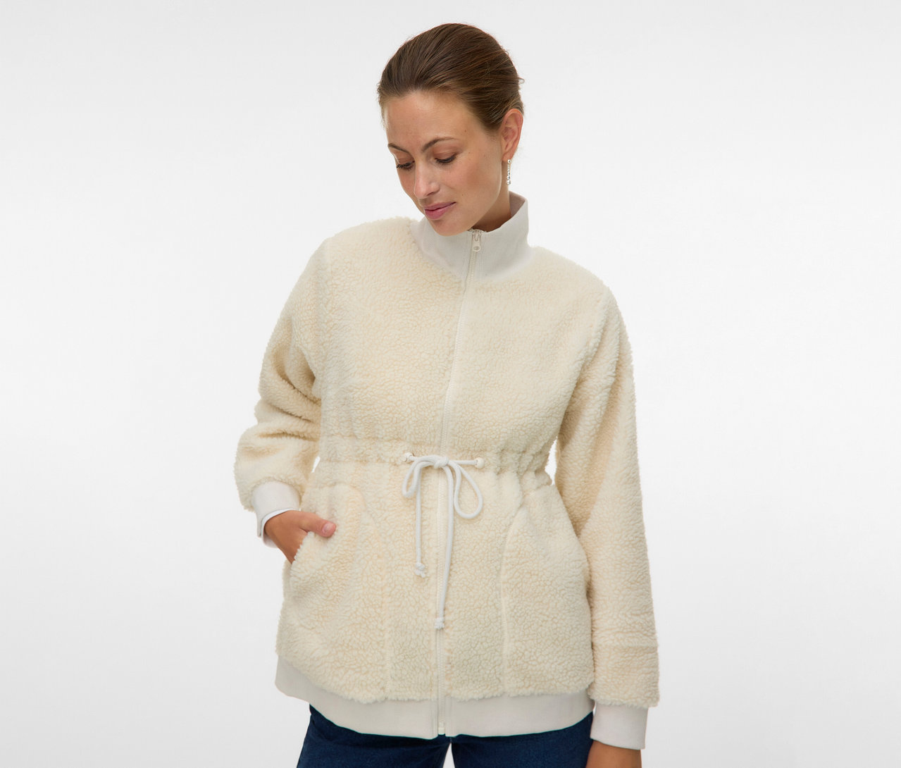 Femme portant la veste Teddy 3 en 1 MAMALICIOUS « MLCARLY » ouverte, de couleur crème, avec cordon de serrage à la taille et mains dans les poches.