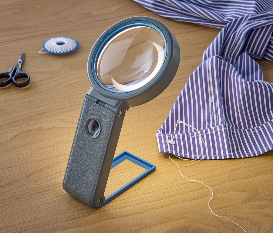Une loupe est posée sur une table en bois à côté de ciseaux, d'une bobine de fil et d'une chemise rayée bleue et blanche.