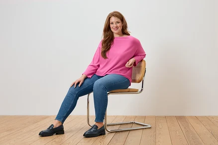 Une femme est assise sur une chaise et porte un pull rose, un jean bleu et des mocassins noirs.