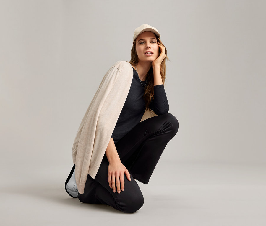 Femme accroupie portant une chemise fonctionnelle outdoor noire, un cardigan beige et une casquette claire.