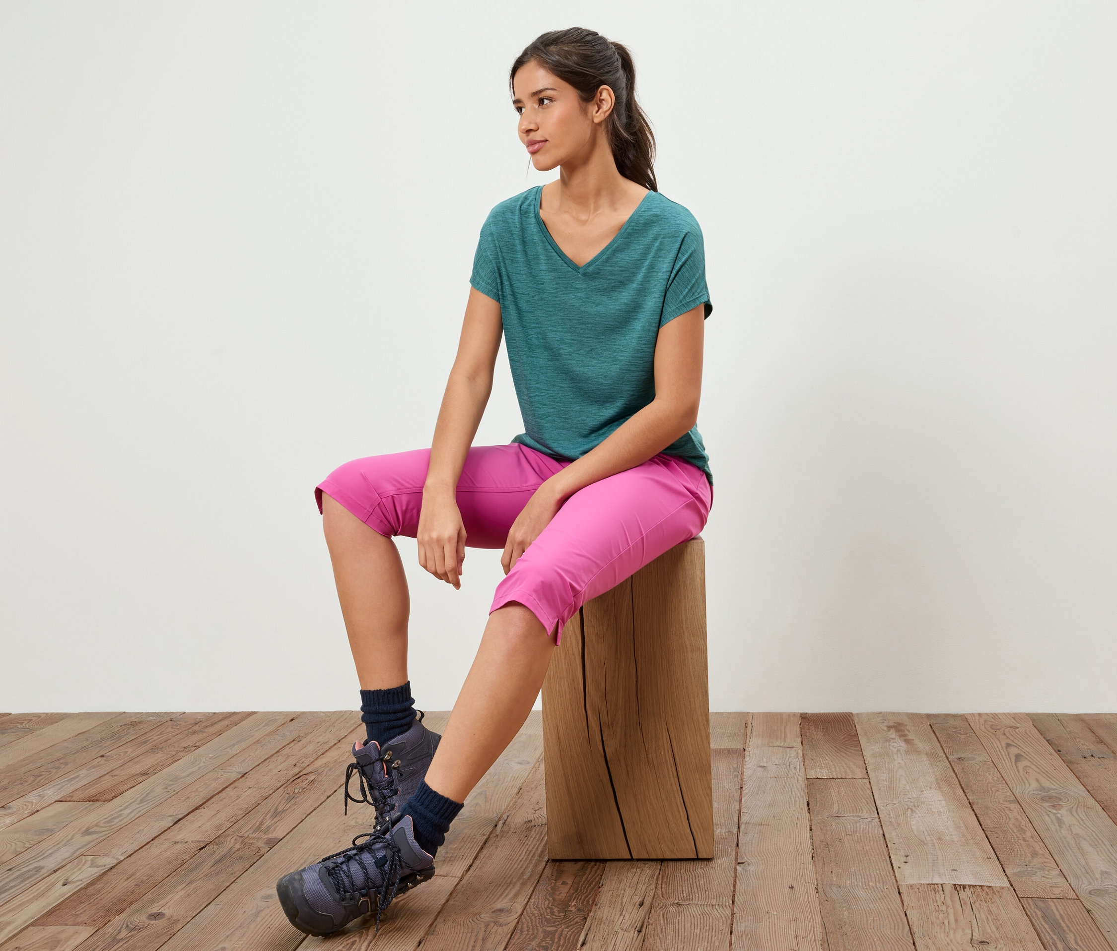 Une femme est assise sur un tabouret en bois, portant un t-shirt vert, un pantacourt rose et des chaussures de randonnée.