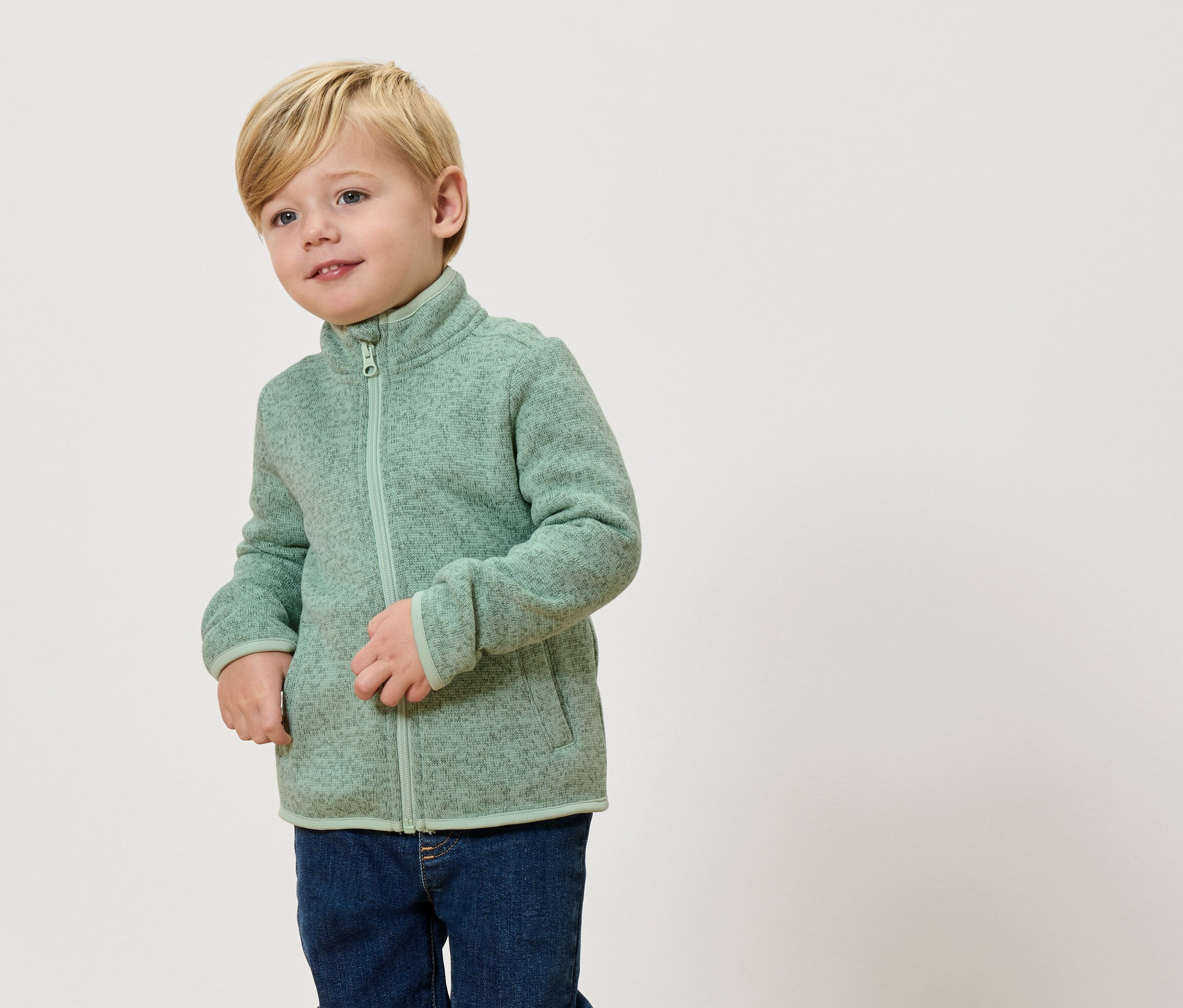 Un garçon blond porte une veste polaire vert clair et un jean.