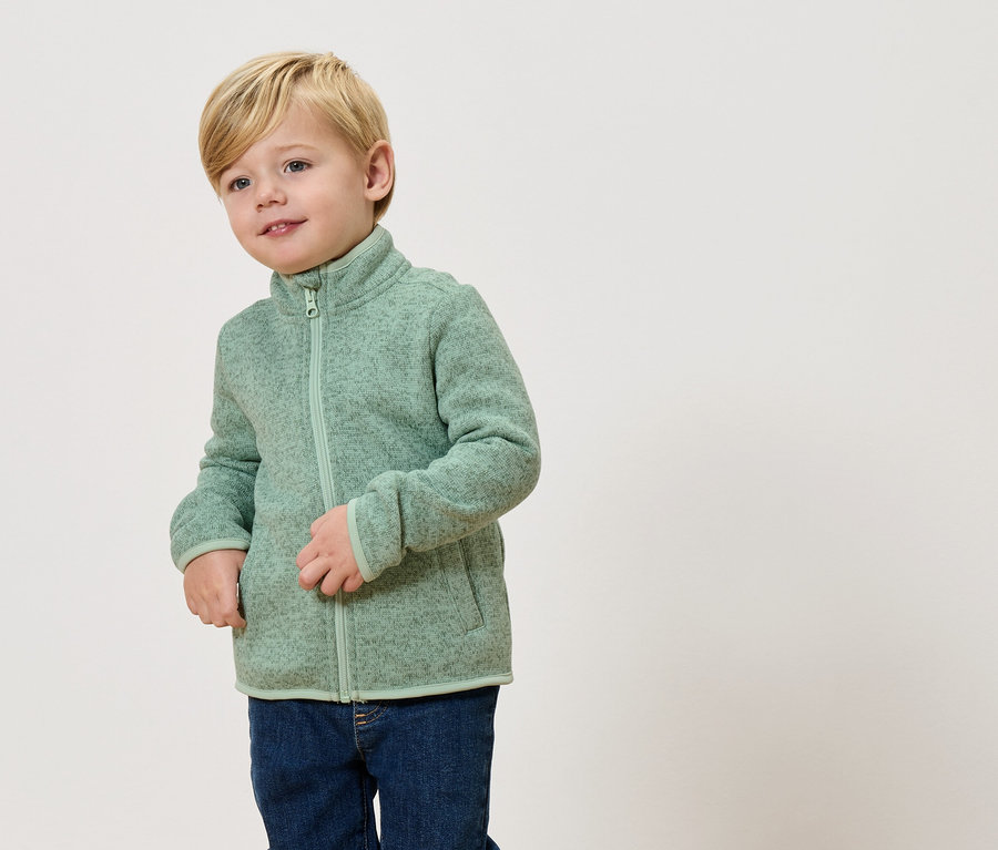 Un garçon blond porte une veste polaire vert clair et un jean.