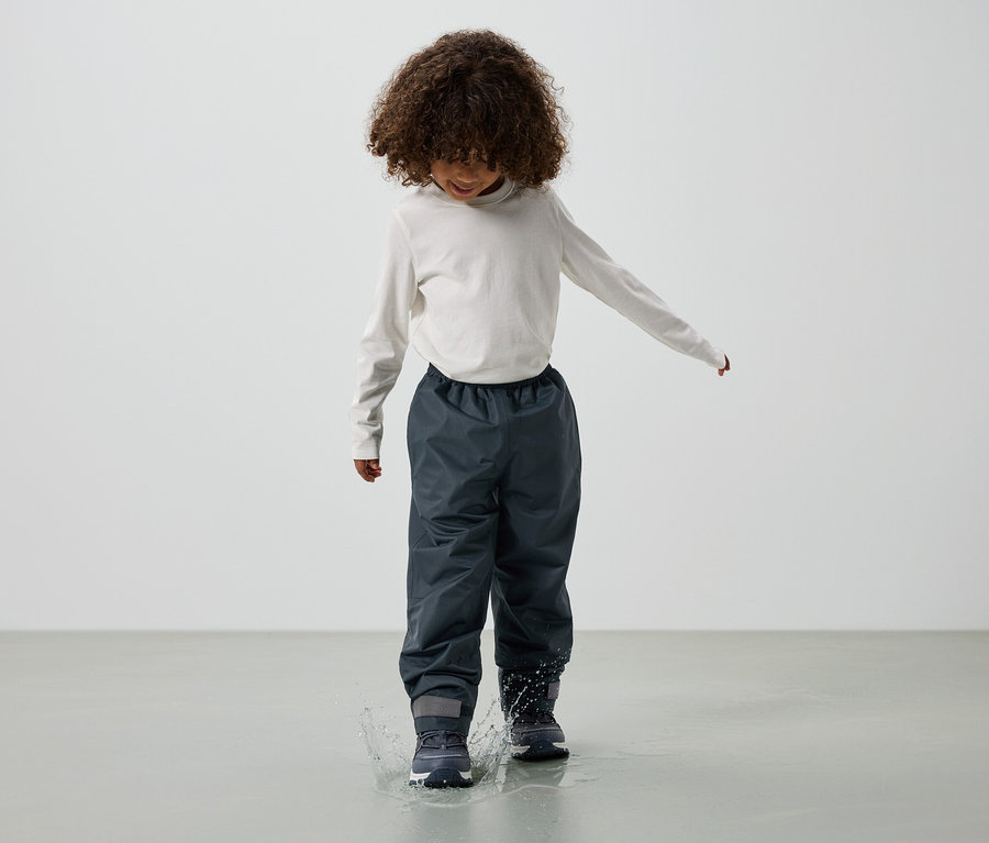 Un enfant aux cheveux bouclés, vêtu d'un haut à manches longues, d'un pantalon de pluie gris et de bottes, saute dans une flaque d'eau.