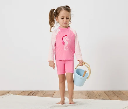 Une petite fille avec des tresses porte un maillot de bain rose avec un motif d'hippocampe et tient un seau bleu sur un plancher en bois.