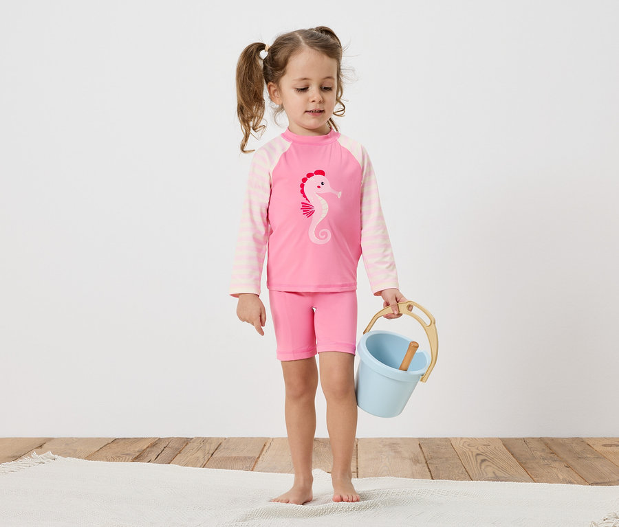 Une petite fille avec des tresses porte un maillot de bain rose avec un motif d'hippocampe et tient un seau bleu sur un plancher en bois.