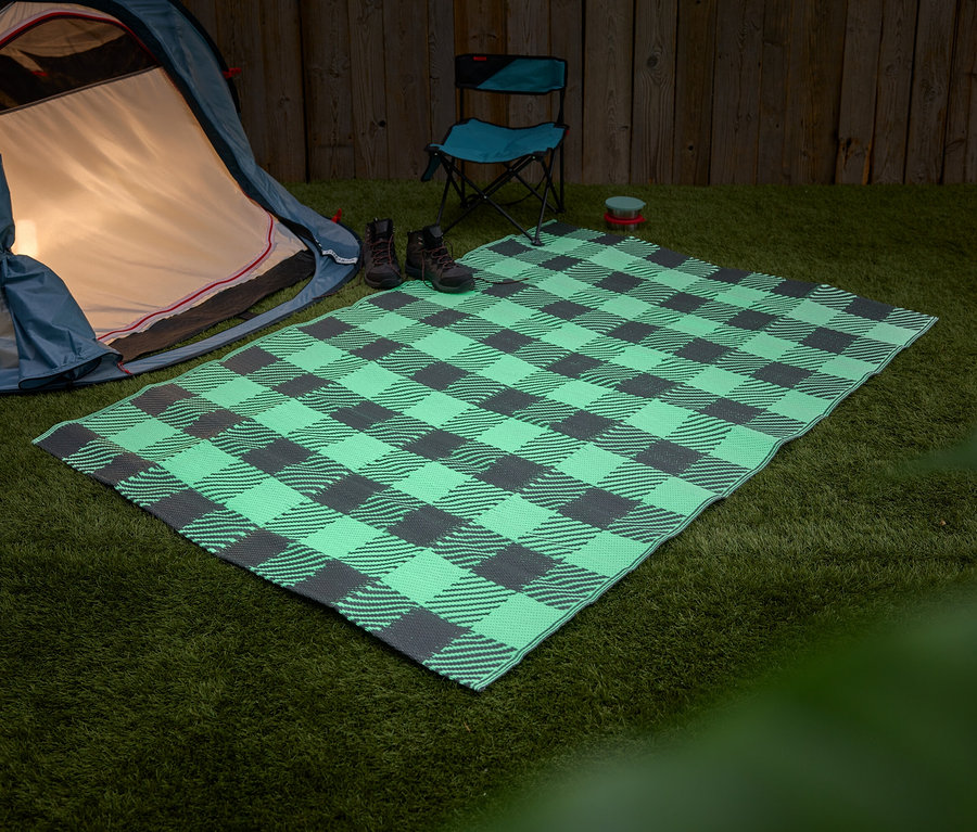 Une tente bleue se dresse à côté d'une couverture de pique-nique à carreaux verts et gris sur de l'herbe. Deux bottes et une chaise pliante bleue sont posées sur la couverture.