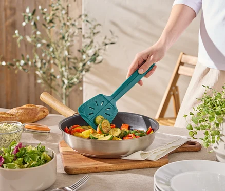 Une personne fait sauter des légumes dans une poêle avec une spatule turquoise. Il y a aussi du pain, de la salade et des herbes sur la table.