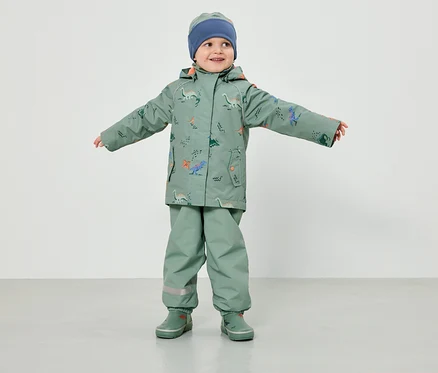 Un jeune enfant portant un vêtement de pluie vert clair à motifs de dinosaures et un bonnet bleu se tient les bras écartés devant un fond blanc.