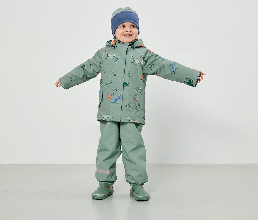 Un jeune enfant portant un vêtement de pluie vert clair à motifs de dinosaures et un bonnet bleu se tient les bras écartés devant un fond blanc.