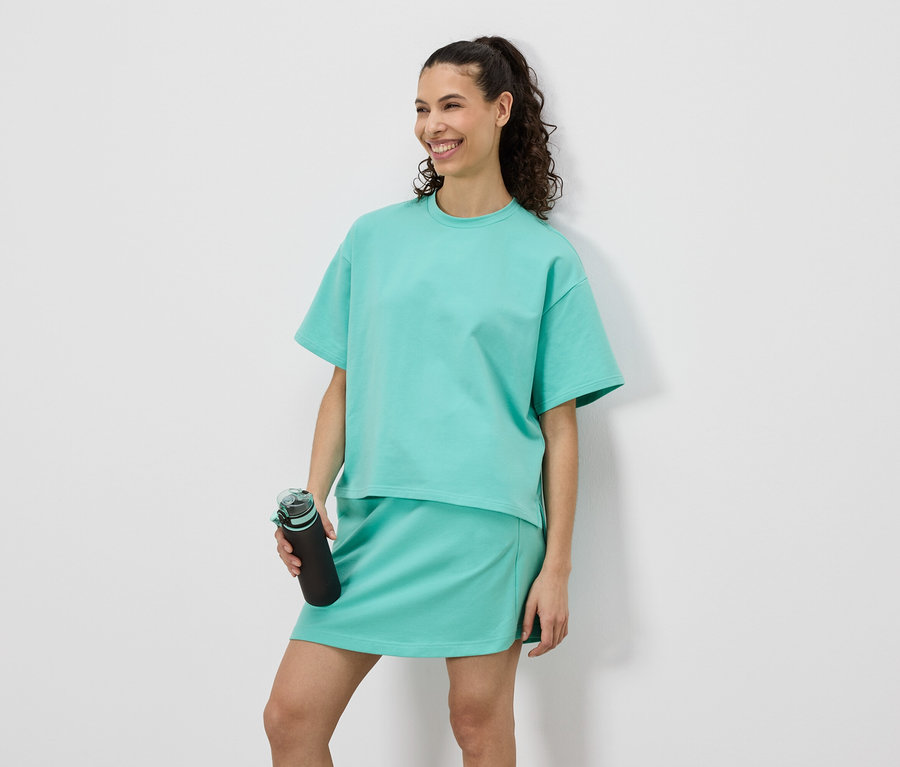 Femme souriante portant un t-shirt et une jupe turquoise tenant une bouteille d'eau noire.