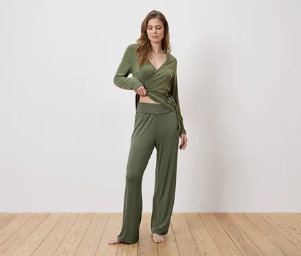 Une femme vêtue d'un haut cache-cœur vert et d'un pantalon se tient pieds nus sur un plancher en bois.