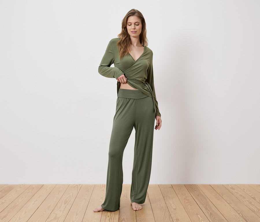 Une femme vêtue d'un haut cache-cœur vert et d'un pantalon se tient pieds nus sur un plancher en bois.