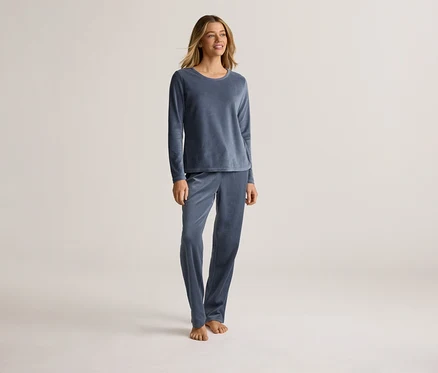 Une femme se tient pieds nus sur un fond clair, vêtue d'un pyjama bleu en velours à manches longues.
