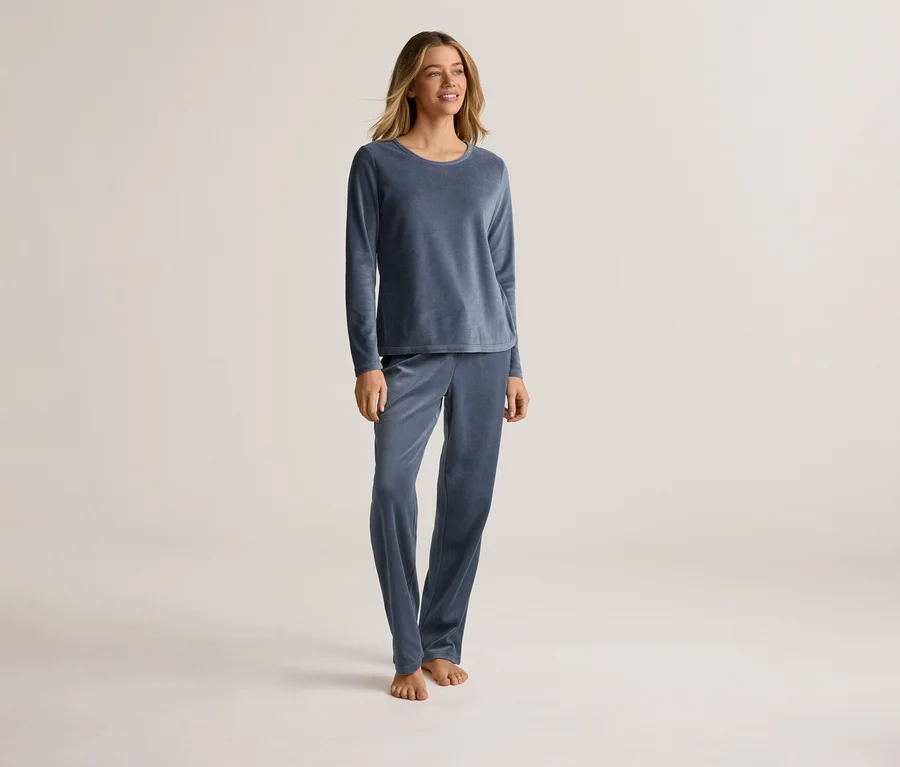 Une femme se tient pieds nus sur un fond clair, vêtue d'un pyjama bleu en velours à manches longues.