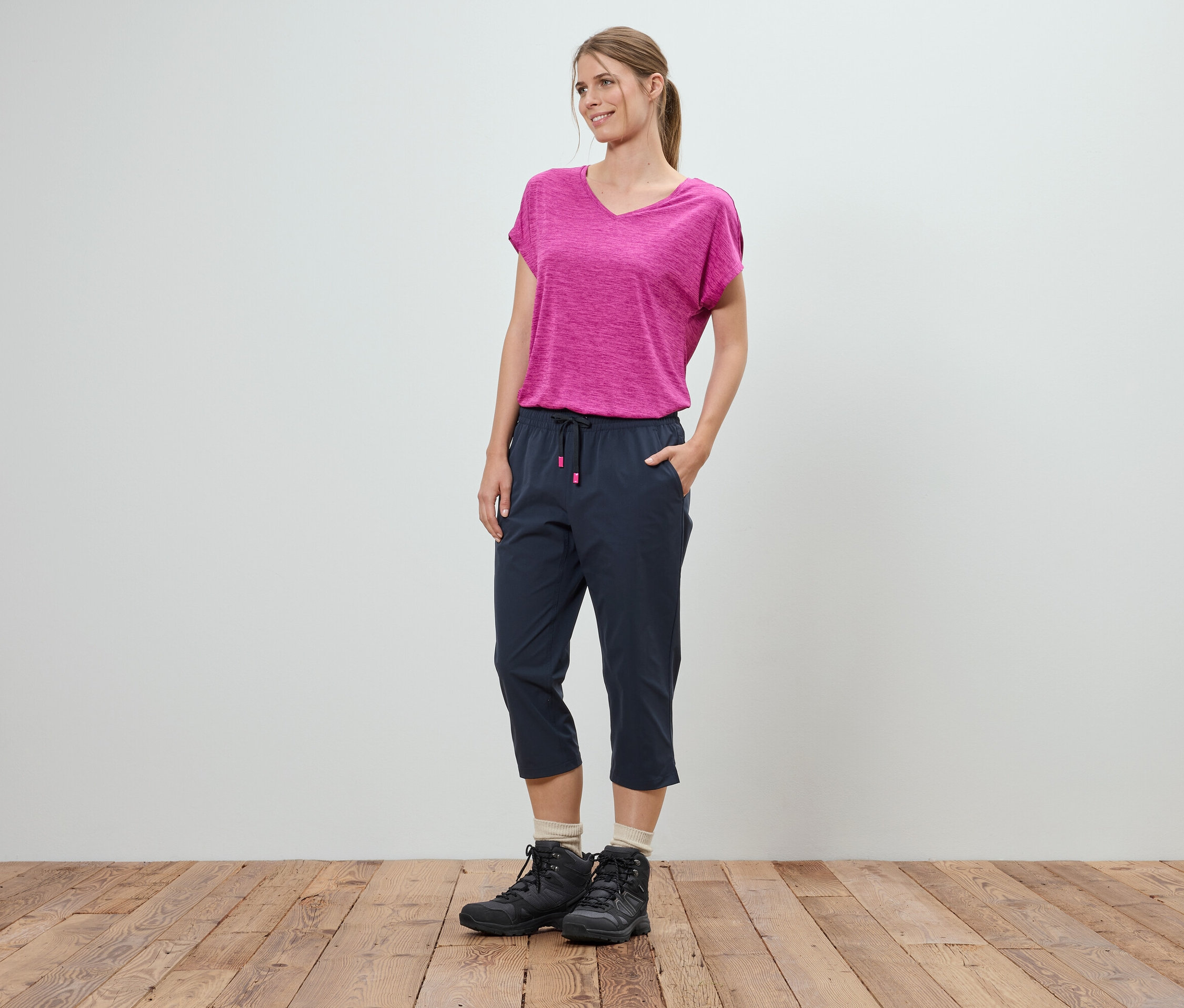Une femme se tient debout, les mains dans les poches, devant un mur blanc. Elle porte un t-shirt rose, un pantacourt bleu et des chaussures de randonnée noires.