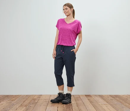 Une femme se tient debout, les mains dans les poches, devant un mur blanc. Elle porte un t-shirt rose, un pantacourt bleu et des chaussures de randonnée noires.