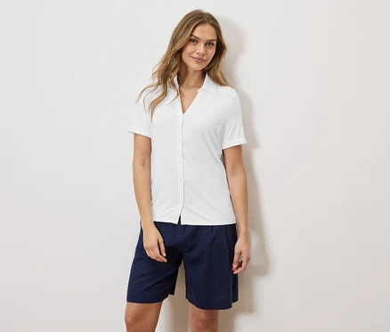 Une femme se tient devant un mur, portant une chemise blanche et un short bleu.