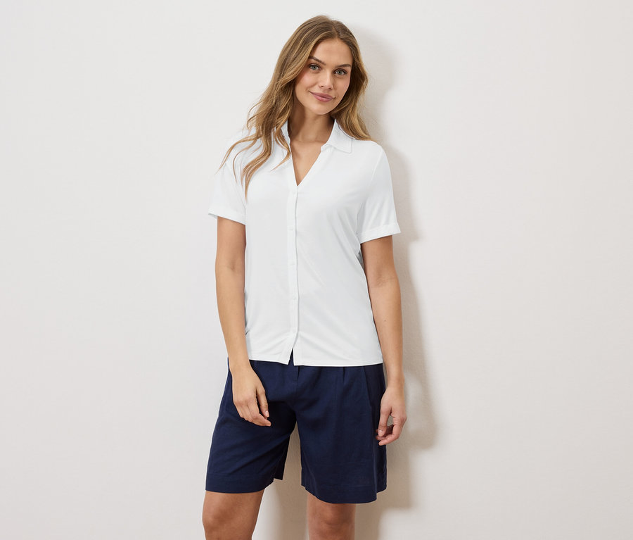 Une femme se tient devant un mur, portant une chemise blanche et un short bleu.