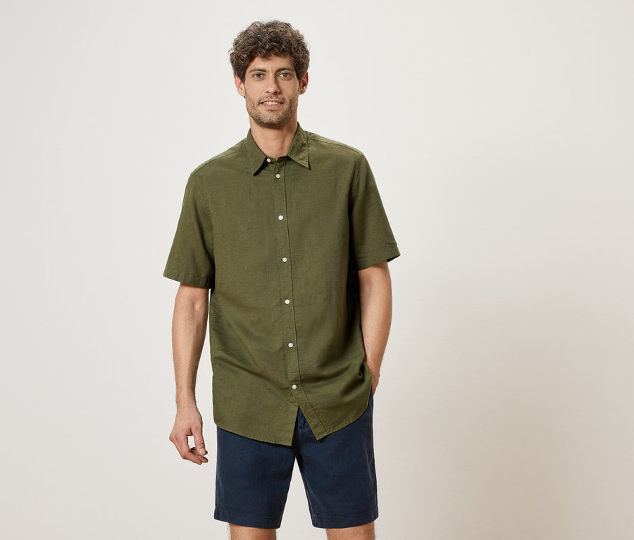 Un homme aux cheveux bouclés se tient debout et porte une chemise vert olive à manches courtes et un short bleu.