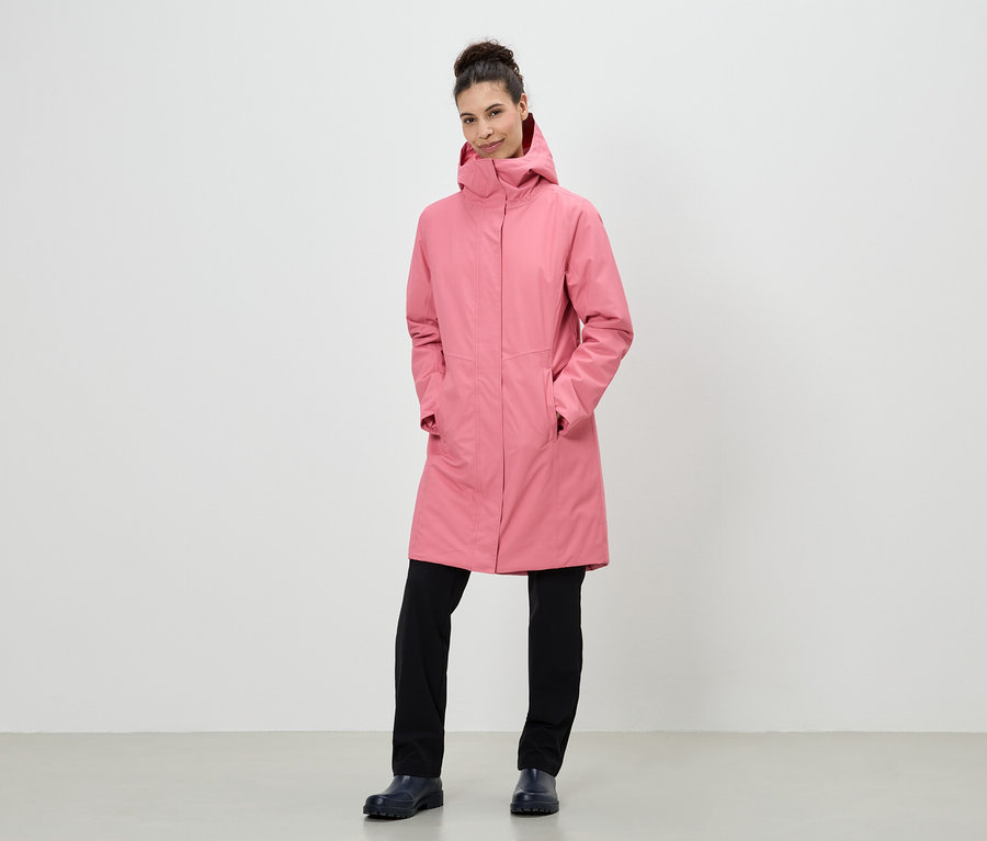 Une femme aux cheveux relevés porte un long manteau rose à capuche, un pantalon noir et des chaussures bleu marine.