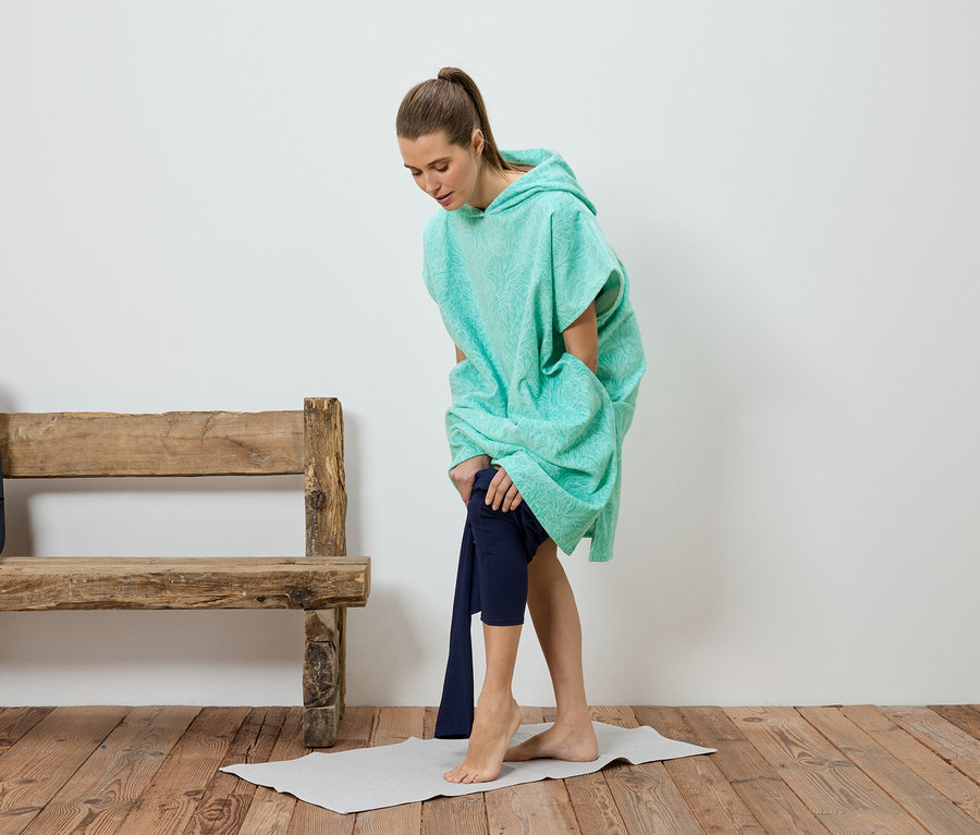 Une femme enfile un pantalon sous un poncho turquoise. Un banc et un sac à dos sont en arrière-plan.