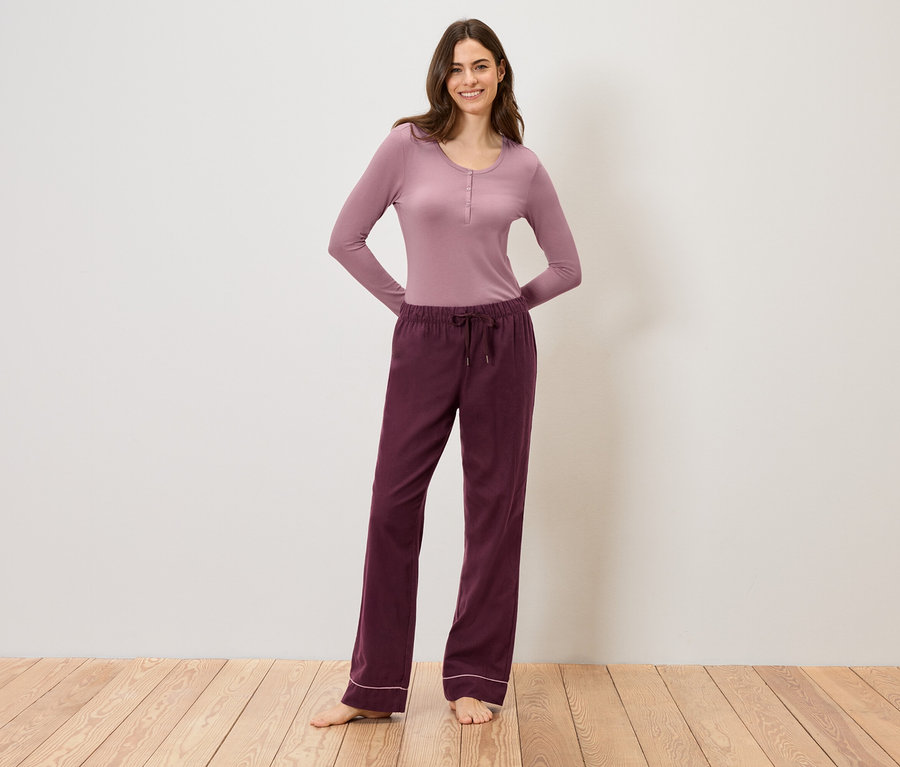 Une femme portant un haut mauve à manches longues et un pantalon bordeaux à cordon de serrage, debout sur un plancher en bois.