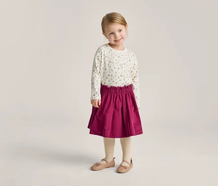 Une petite fille blonde porte un haut à manches longues à fleurs, une jupe rouge, des collants et des ballerines.