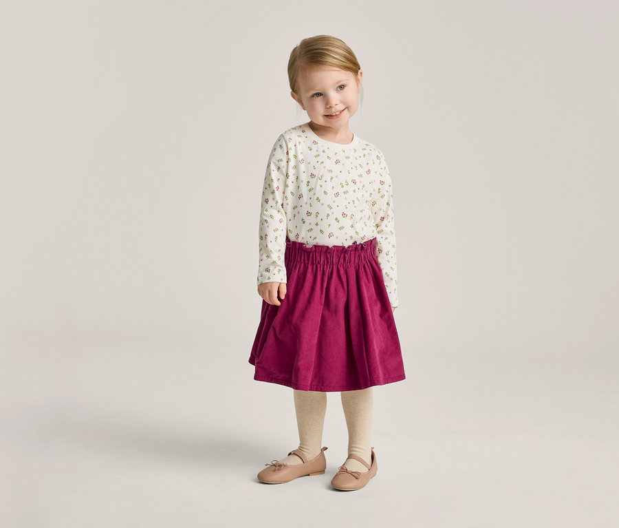 Une petite fille blonde porte un haut à manches longues à fleurs, une jupe rouge, des collants et des ballerines.