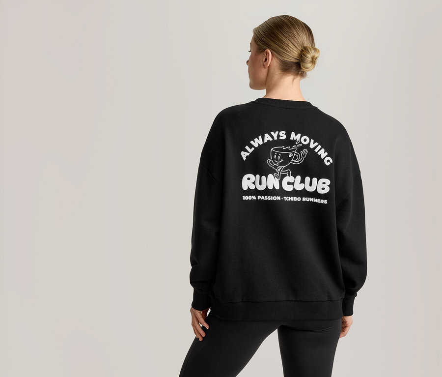 Une femme vue de dos porte un Sweatshirt noir avec un motif blanc au dos. Le motif représente un personnage de tasse de café de dessin animé et le texte 'ALWAYS MOVING RUN CLUB' et '100% PASSION - TCHIBO RUNNERS'.