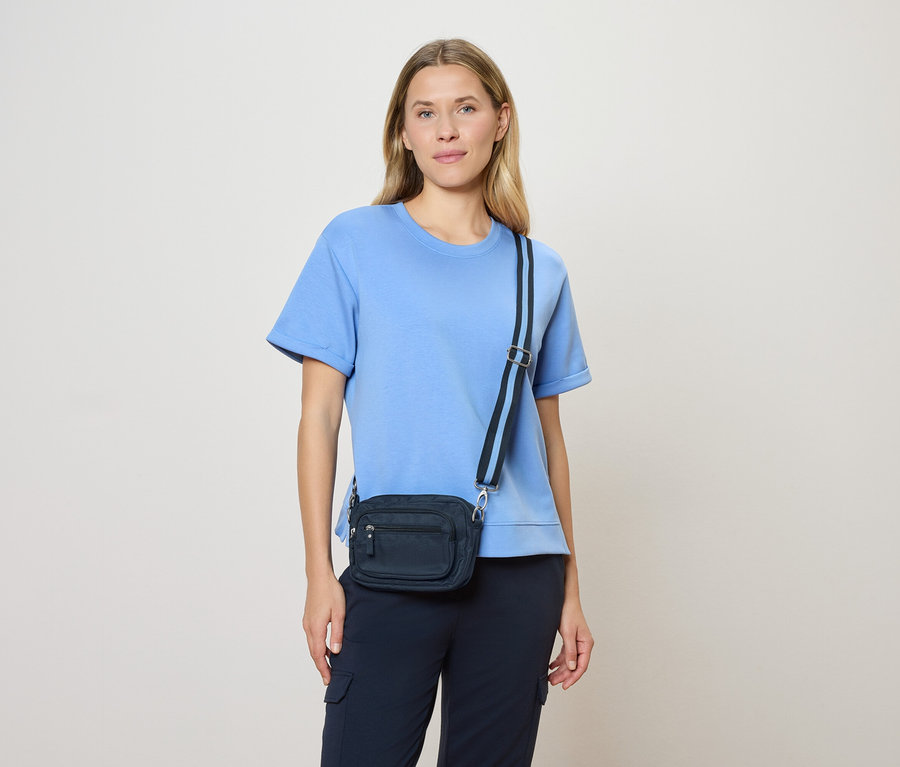 Femme aux cheveux châtain clair portant un t-shirt bleu clair et un sac à bandoulière bleu marine avec une bandoulière bleu marine devant un fond crème.
