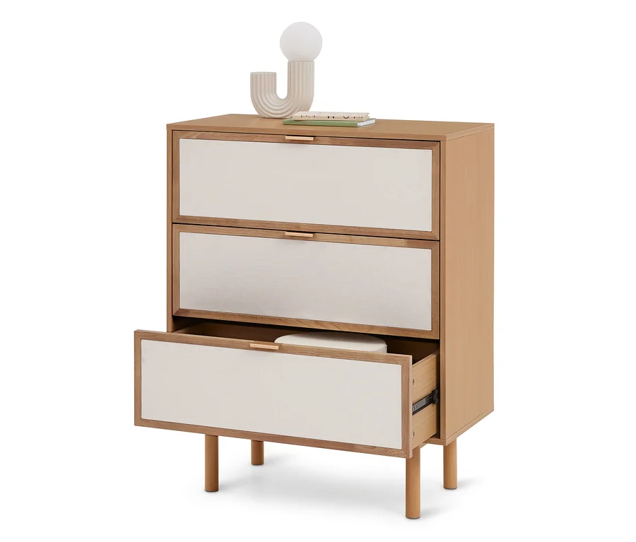 Commode en bois clair avec trois tiroirs à façade en tissu crème et poignées dorées. Le tiroir inférieur est ouvert, révélant un objet blanc. Une lampe décorative et des livres sont posés dessus.