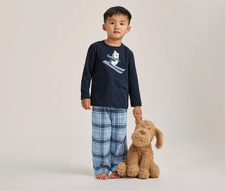 Un jeune garçon pieds nus porte un pyjama bleu marine à manches longues avec un motif de pingouin skieur et un pantalon à carreaux bleus, tenant un chien en peluche marron.