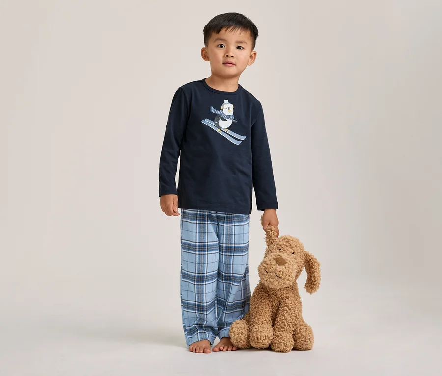 Un jeune garçon pieds nus porte un pyjama bleu marine à manches longues avec un motif de pingouin skieur et un pantalon à carreaux bleus, tenant un chien en peluche marron.